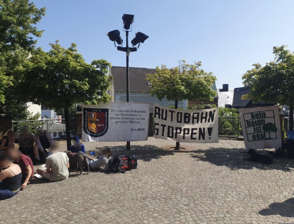 Transpis und Picknickrunde im Sonnenschein vor dem Landgericht Flensburg. Auf den drei Transpis steht: "Kein Baum ist Egal", "Autobahn stoppen!" und "Mobile Einsatzgruppe Justiz: Wir treten mit Schlagstock und Handschellen in blindem Gehorsam für eine gerechtere Welt ein"